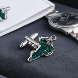 May include: Silver cufflinks featuring a green leaf design, alongside a white shirt cuff and a silver wristwatch. The cufflinks have a detailed, nature-inspired aesthetic, with a dark green enamel leaf set in a silver frame.