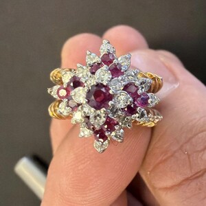 Può includere: Un anello a grappolo con un rubino centrale rosso intenso dal taglio rotondo circondato da rubini più piccoli e diamanti bianchi chiari incastonati in metallo argentato, montato su una fascia in oro giallo testurizzato a corda.
