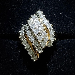 May include: A gold ring featuring a cluster of numerous small, round, clear, faceted stones arranged in a diagonal, layered, wave-like pattern across the top. The stones are set closely together, creating a dense, sparkling surface texture.