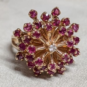 Puede incluir: Un anillo de oro con un grupo de numerosos rubíes redondos de color rosa intenso dispuestos en forma de estrella. Un solo diamante pequeño, transparente y de talla redonda está engastado en el centro exacto, rodeado por los radios con rubíes.