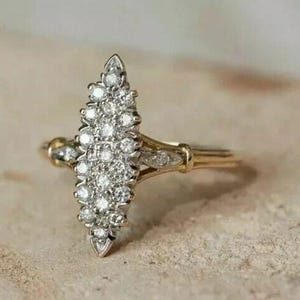 Peut inclure: Une bague de forme marquise en or jaune, ornée d'un groupe central de nombreuses petites pierres blanches rondes taille brillant serties dans du métal blanc. L'anneau est fin et poli, avec un petit diamant accentué de chaque côté.