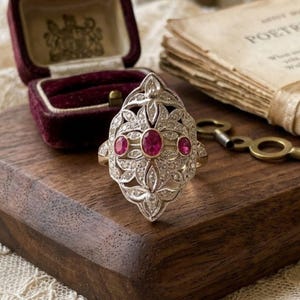 Puede incluir: Un anillo de tono plateado con un diseño de filigrana alargado e intrincado, engastado con tres piedras preciosas ovaladas de color rojo rosado vibrante dispuestas horizontalmente en el centro, rodeadas de pequeñas piedras brillantes.