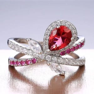 May include: A silver-toned ring featuring a large pear-shaped red centre stone surrounded by a halo of small clear stones. The band has a crossover design encrusted with alternating rows of small clear stones and small round red stones.