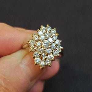 May include: A gold ring with a cluster of clear, round gemstones. The gemstones are arranged in a floral pattern, creating a sparkling effect. The ring is held against a dark background.