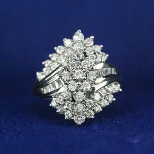 May include: A silver-toned ring featuring a cluster of numerous round, clear, faceted stones arranged in a dense, multi-layered floral pattern. The band is polished and smooth, supporting the central arrangement of sparkling, reflective stones.