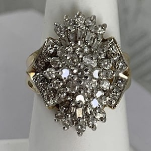Op de afbeelding: Een gouden ring met een grote, ovaalvormige cluster van talloze kleine, ronde en baguette-geslepen diamanten gezet in wit metaal. De centrale opstelling is omringd door extra diamantaccenten, wat zorgt voor een dicht, veelzijdig en reflecterend oppervlak bovenop de ringband.