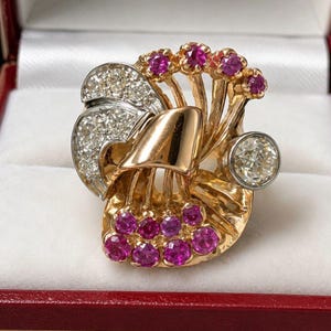 May include: A gold ring featuring a central polished gold ribbon motif, accented with a cluster of round faceted pink rubies at the base and top, a fan-shaped section of pavé-set clear diamonds, and a single large round clear diamond in a bezel setting.