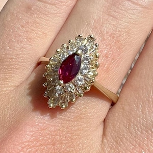 Peut inclure: Une bague en or avec un rubis rouge profond taille marquise au centre, entouré d'un groupe de petites pierres claires taille baguette et ronde disposées en forme ovale allongée et pointue, sur un anneau en or fin et poli.