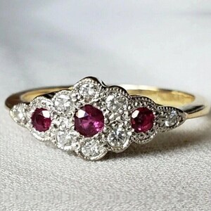 May include: A gold ring featuring a cluster setting with three round red rubies and ten small round white diamonds. The stones are arranged in a horizontal floral-like pattern with milgrain detailing along the edges of the metal setting.