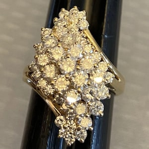 Peut inclure: Une bague en or ornée d'un groupe de nombreuses pierres rondes, transparentes et facettées, disposées dans un sertissage dense en forme de marquise allongée. Les pierres sont serrées, couvrant toute la surface supérieure de l'anneau en or.