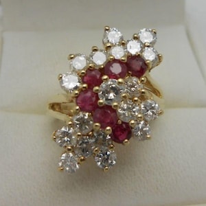 Peut inclure: Une bague en or ornée d'un groupe de rubis rouges et de diamants blancs transparents taille ronde disposés en motif floral. Les pierres précieuses sont serties sur un anneau en or jaune, créant un éclat dense aux couleurs contrastées.