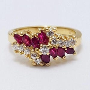 Op de afbeelding: Een gouden ring met een cluster van dieprode robijnen in marquise-slijpvorm en heldere diamanten in ronde briljant-slijpvorm. De stenen zijn gerangschikt in een dicht, meerlaags bloemachtig patroon op een gepolijste geelgouden band.