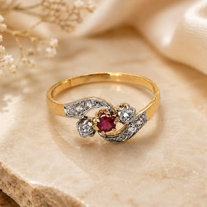 Puede incluir: Un anillo de oro con un rubí rojo redondo central flanqueado por dos pequeños diamantes redondos transparentes. El engaste está hecho de una combinación de una banda de oro amarillo y una montura decorativa de metal blanco con curvas.