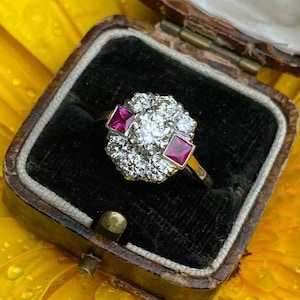 May include: A ring featuring a central round brilliant-cut diamond surrounded by a cluster of smaller round diamonds and two square-cut red rubies set in a metal band, presented inside a dark-lined, hinged jewellery box with a wooden exterior.