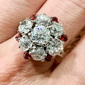 May include: A cluster ring featuring a large central round-cut clear stone surrounded by a halo of smaller round-cut clear stones and interspersed with small, vibrant red round-cut stones, all set in a metallic band.