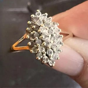 Peut inclure: Une bague en or ornée d'un groupe de nombreuses petites pierres rondes et transparentes disposées en forme de marquise. L'anneau est fin et métallique, soutenant la disposition dense de pierres facettées créant une surface brillante.