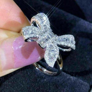 May include: A silver-toned ring featuring a prominent bow-shaped centrepiece. The bow is densely encrusted with numerous small, rectangular baguette-cut stones and smaller round stones, creating a highly reflective and sparkling surface.