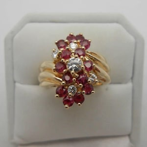 Peut inclure: Une bague en or ornée d'un groupe de rubis rouges ronds et de diamants blancs transparents disposés en motif floral. L'anneau se compose de plusieurs brins d'or lisses et polis qui convergent pour soutenir le sertissage des pierres.