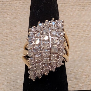 Puede incluir: Un anillo de oro con una cabeza de racimo ancha de varias filas, densamente engastada con numerosas piedras pequeñas, redondas, claras y facetadas. La banda se compone de varios hilos de oro paralelos que convergen en el engaste principal.