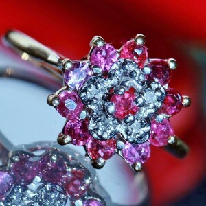 Peut inclure: Une bague avec un sertissage en forme de fleur en métal argenté. Le centre est orné d'une pierre rose ronde entourée d'un cercle de petites pierres claires et brillantes, avec une bordure extérieure de dix pierres roses rondes.