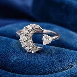 Puede incluir: Un anillo abierto de tono plateado que presenta un motivo de media luna incrustado con pequeñas piedras claras y una piedra clara única en forma de corazón en el extremo opuesto. El anillo tiene un acabado metálico pulido y un diseño abierto.