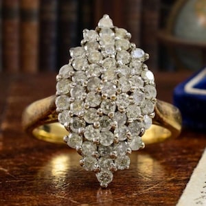 May include: A gold ring featuring a large, marquise-shaped cluster head composed of numerous small, round, clear stones set closely together. The band is smooth and metallic gold, supporting the prominent, elongated, multi-stone centerpiece.
