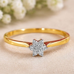 Peut inclure: Une bague en or jaune avec un sertissage en forme de fleur au centre. Le motif floral se compose de six petites pierres rondes et transparentes disposées autour d'une pierre centrale, le tout monté sur une monture argentée et un anneau lisse.