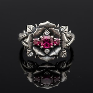 May include: A silver-toned ring featuring a rose design with a large, square, magenta-colored gemstone at the center, flanked by two smaller gems. The rose is surrounded by leaves and small, clear accent stones.