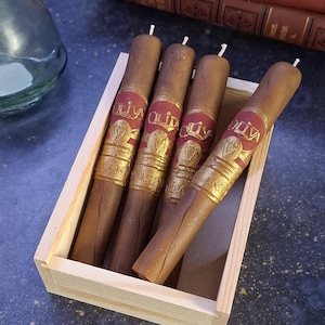 May include: Four brown cigar-shaped candles with red and gold labels, presented in a light wooden box. The candles have white wicks and are arranged at an angle. The labels read "OLIVA".