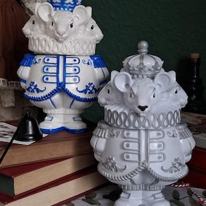 Può includere: Due statuine decorative di topi bianchi con più teste, che indossano abiti regali. Una statuina ha dettagli blu, l'altra argentati. Entrambe hanno corone e sono in piedi. Le statuine sono su un tavolo con libri.