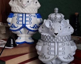 Puede incluir: Dos figuras decorativas de ratones blancos con múltiples cabezas, vestidas con atuendo real. Una figura tiene detalles azules, la otra plateados. Ambas tienen coronas y están de pie. Las figuras están sobre una mesa con libros.