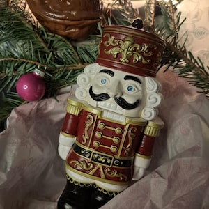 Può includere: Statuina decorativa schiaccianoci in un'ambientazione festosa. La statuina indossa un'uniforme rossa e oro con cappello e stivali neri. Ha un viso bianco con occhi blu e baffi neri. Lo sfondo comprende rami e ornamenti dell'albero di Natale.