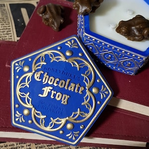May include: A blue and gold Honeydukes Chocolate Frog box with the text "Chocolate Frog" and a frog-shaped candle. The box is open, revealing a white candle and three frog-shaped decorations. The box is on top of a red book.