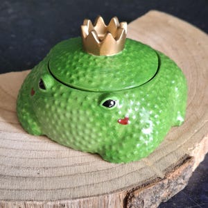 Könnte beinhalten: Grüne, froschförmige Schmuckdose aus Keramik mit goldener Krone. Der Frosch hat eine strukturierte Oberfläche, schwarze Augen und rote Lippen. Die Dose liegt auf einer Holzscheibe.