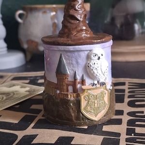 May include: A decorative jar with a brown sorting hat lid. The jar features a Hogwarts crest, a castle, and an owl. The jar is painted in shades of purple, brown, and gold.