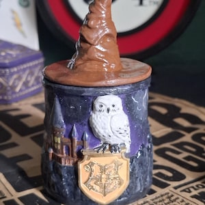 Puede incluir: Un recipiente cilíndrico con una tapa en forma de sombrero marrón arrugado. El exterior presenta un fondo de cielo nocturno morado y negro, un castillo marrón detallado, un búho blanco sobre un emblema dorado y un escudo dorado.