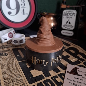Può includere: Un portagioie marrone e nero a tema Harry Potter Sorting Hat. La scatola ha le parole "Harry Potter" in lettere dorate. Il Sorting Hat è in cima alla scatola. Altri oggetti a tema Harry Potter sono sullo sfondo.