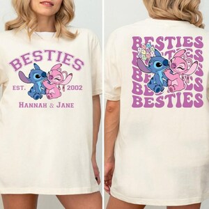 Puede incluir: Camiseta color crema con un dise&ntilde;o de personajes de dibujos animados azules y rosas y la palabra "BESTIES" en un arco morado. La parte delantera de la camiseta tiene el texto "EST. 2002" y los nombres "Hannah & Jane". La parte trasera tiene el mismo dise&ntilde;o.