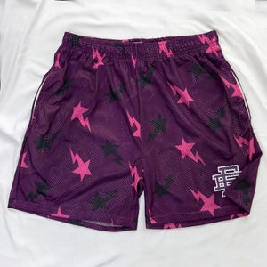 May include: Purple mesh shorts featuring a pattern of pink lightning bolts and stars, with some black stars. The shorts have white trim on the sides and a white logo on the right leg.