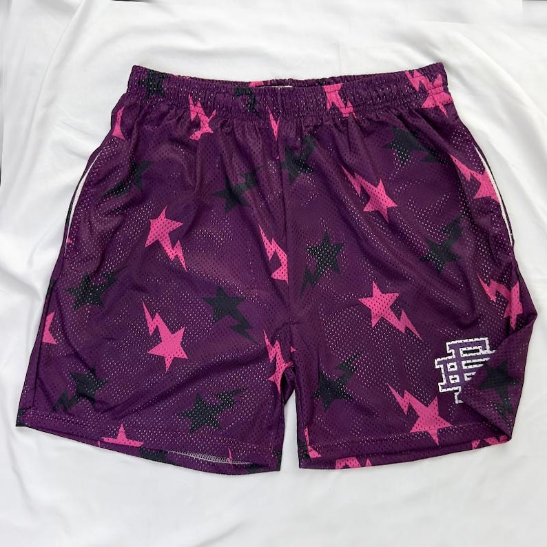 May include: Purple mesh shorts featuring a pattern of pink lightning bolts and stars, with some black stars. The shorts have white trim on the sides and a white logo on the right leg.