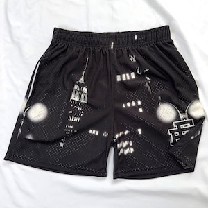 May include: Black athletic shorts featuring a white graphic print of a city skyline at night, including a tall skyscraper and illuminated windows. The shorts are made of a perforated mesh fabric and have an elastic waistband for a secure fit.