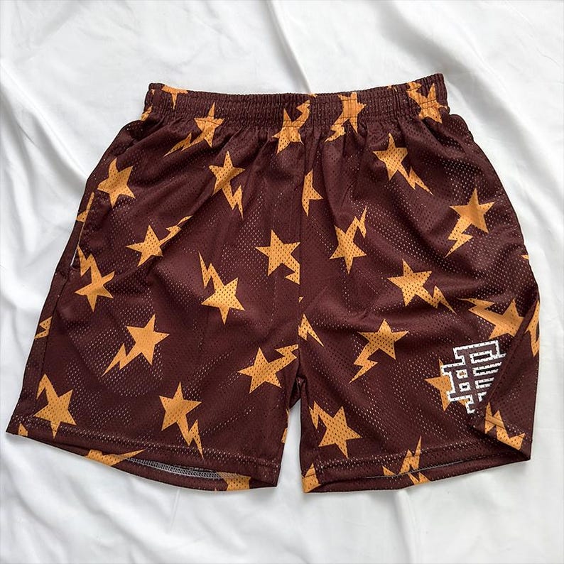 May include: Brown mesh shorts featuring a repeating pattern of gold stars and lightning bolts. The shorts have an elasticated waistband and a white logo on the lower right leg. The fabric appears lightweight and breathable.