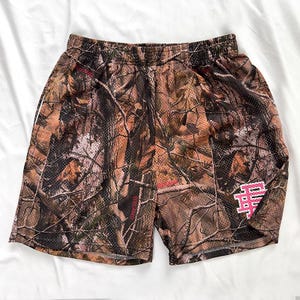 May include: Camouflage print mesh shorts in shades of brown, green, and tan. The shorts feature a pink logo on the lower right leg and pink text. The shorts have an elasticated waistband.