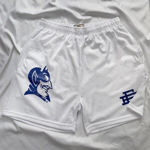 May include: White mesh shorts featuring a blue devil logo and a blue stylized letter logo. The shorts have an elasticated waistband. The shorts are designed for sports.
