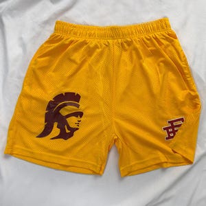 May include: Yellow mesh shorts featuring a maroon gladiator helmet graphic on the left leg and a maroon letter graphic on the right leg. The shorts have an elasticated waistband.