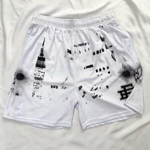 May include: White mesh athletic shorts with a black graphic print. The shorts have an elasticated waistband and a black logo on the lower right leg. The graphic appears to be a stylized cityscape.