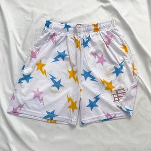 May include: White mesh shorts with an elasticated waistband, patterned with blue, yellow, and pink stars and lightning bolts. A small logo is on the right leg. Casual athletic wear.