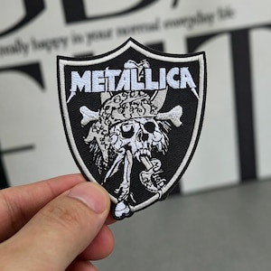 Pode incluir: Patch bordado em preto e branco com a palavra "METALLICA" em letras brancas. O patch apresenta uma caveira com ossos cruzados e uma forma de escudo. O patch é segurado por uma mão.