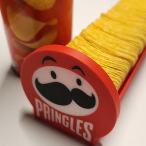 Könnte beinhalten: Eine rote Pringles-Dose und ein roter Chiphalter mit einem Stapel gelber Chips. Der Chiphalter zeigt das Pringles-Logo mit Schnurrbart und dem Wort "PRINGLES" in Weiß.
