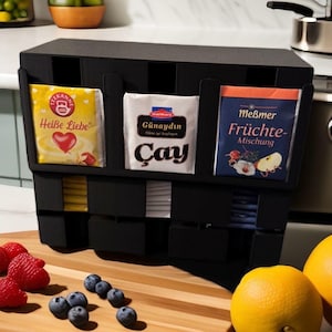 Könnte beinhalten: Ein schwarzer Teebeutel-Organizer mit drei Fächern, die jeweils verschiedene Teebeutel enthalten. Die Beutel sind in gelben, weißen und blauen Verpackungen. Frische Früchte, darunter Erdbeeren, Blaubeeren und Zitronen, befinden sich im Vordergrund.